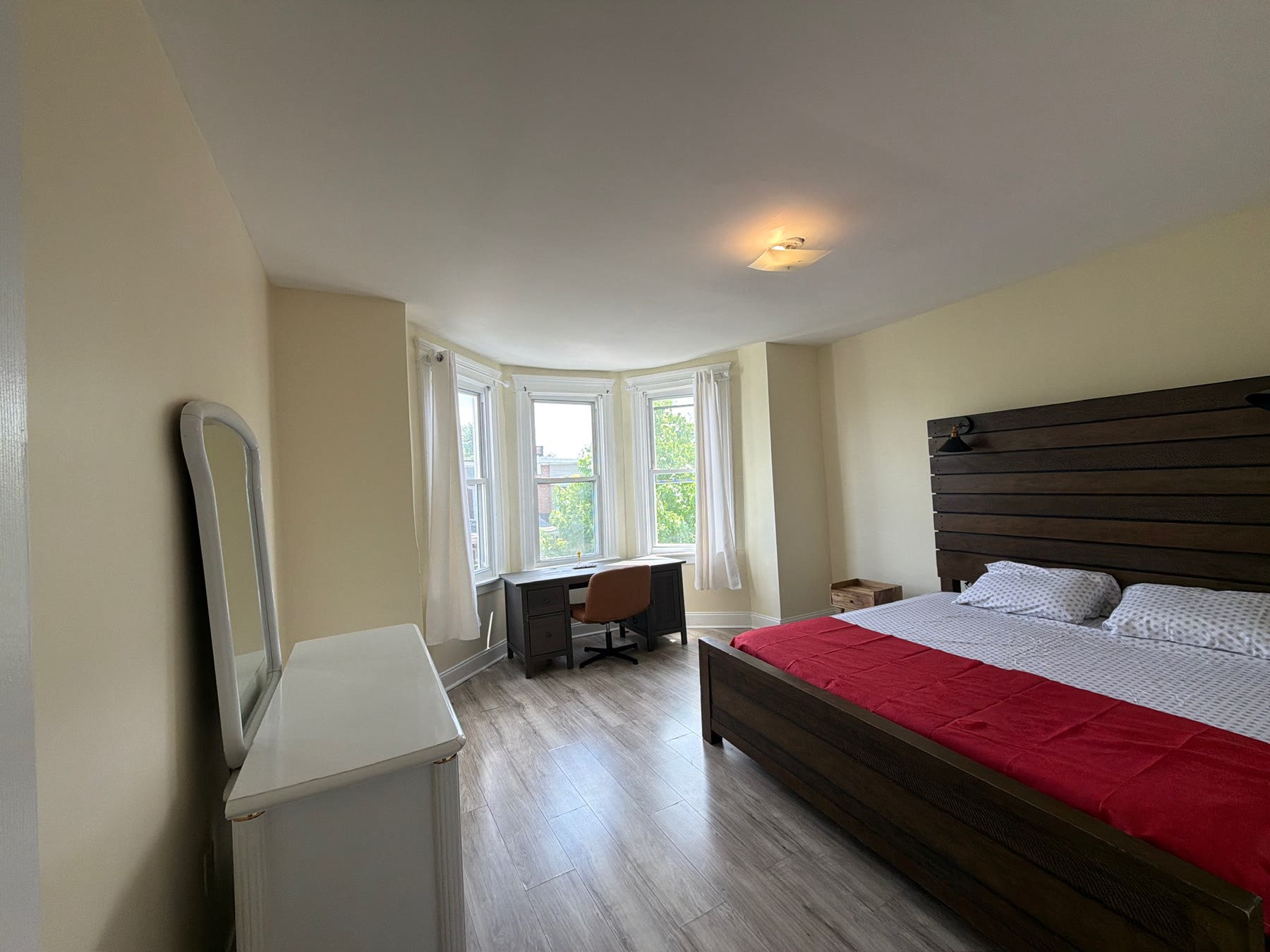 Room 203 at Leia: a wide bed with a dark slatted headboard and a red throw, in a bay-windowed room with a desk and a tall standing mirror.