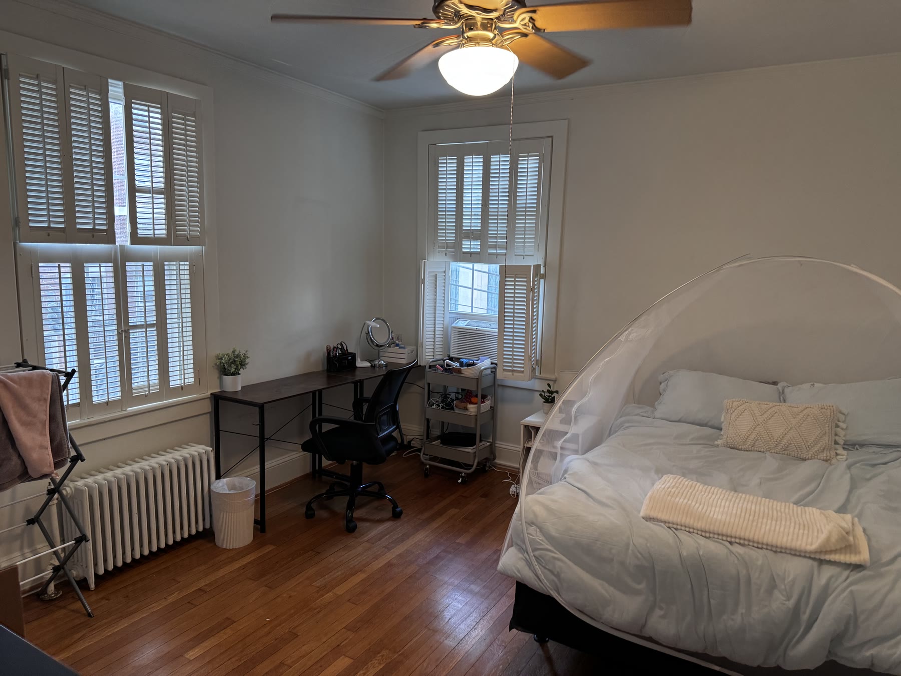 Room 102 at Lincoln F: a bed and a desk under shuttered windows on three sides, with dark wood floors and a ceiling fan.