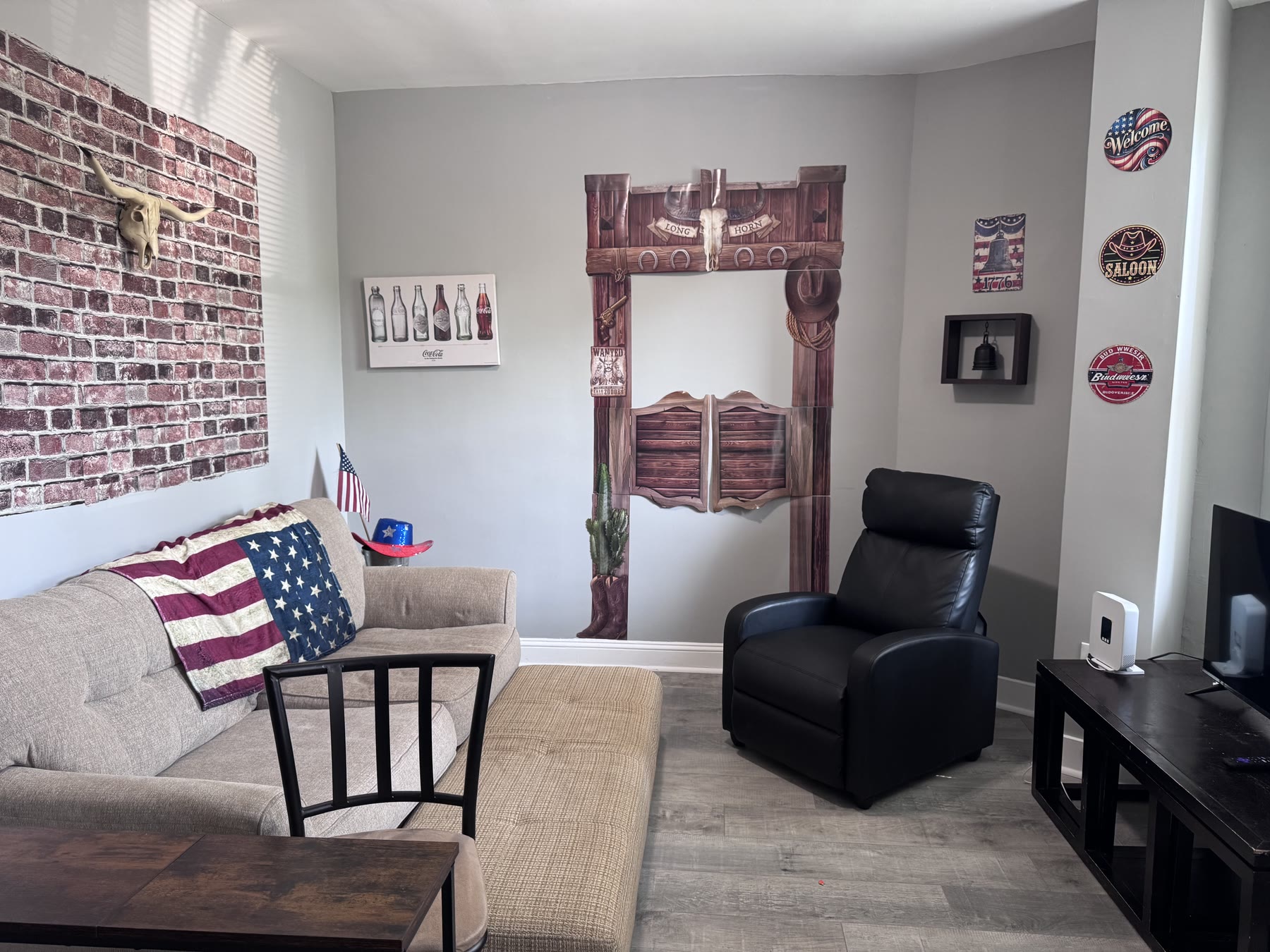 Mohica's living room: a sofa and recliner facing the television, with saloon doors and a brick wall hung with stars-and-stripes cushions and road signs.