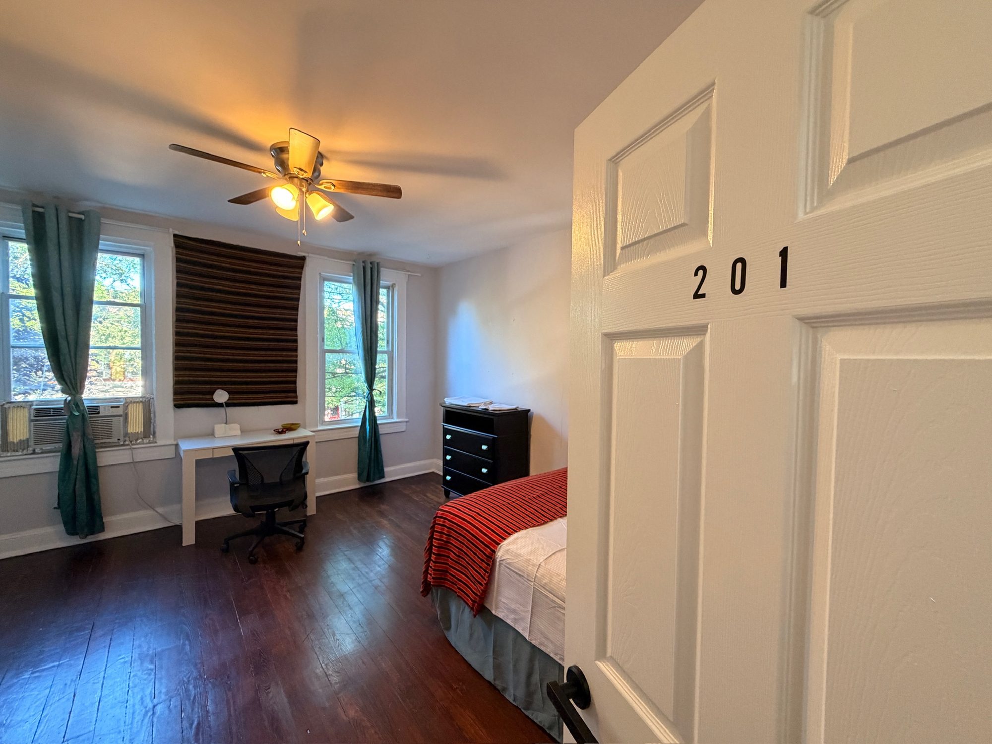Room 201 at Ujamaa: a bed with a red throw and a desk under curtained windows, seen past the numbered door on dark wood floors.
