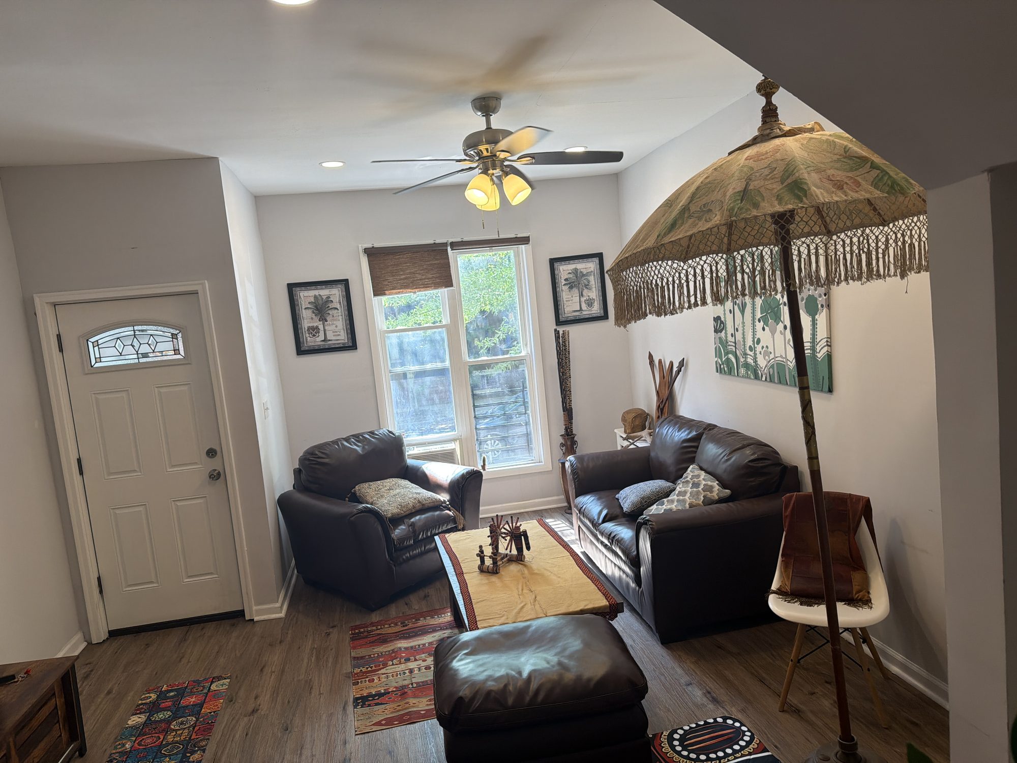 Ujamaa's living room: leather sofas and a kilim rug around a low table, with a fringed lamp and framed pictures beside the front door.